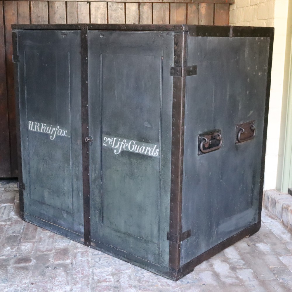 A large dark green metal-bound military campaign travelling case with stencilled lettering on the front panels and iron drop handles on the side.