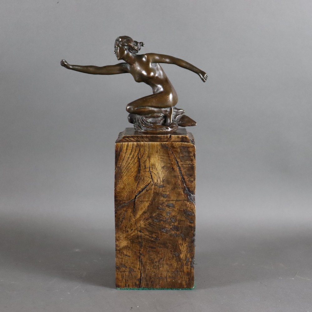 An Art Deco dark bronze sculpture of a kneeling nude woman reaching forward, mounted on a rustic square timber base.