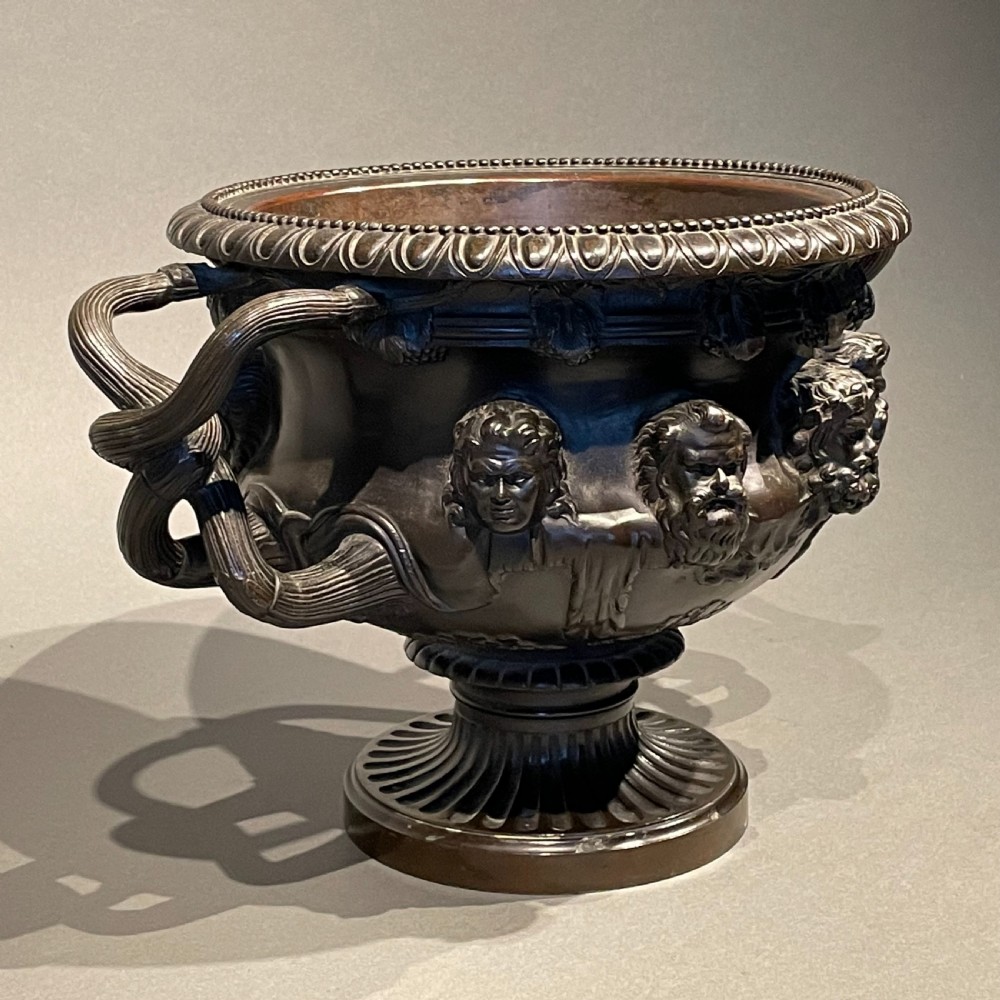A dark patinated bronze Warwick vase featuring relief Bacchic masks, intertwined vine handles, and a gadrooned pedestal foot.
