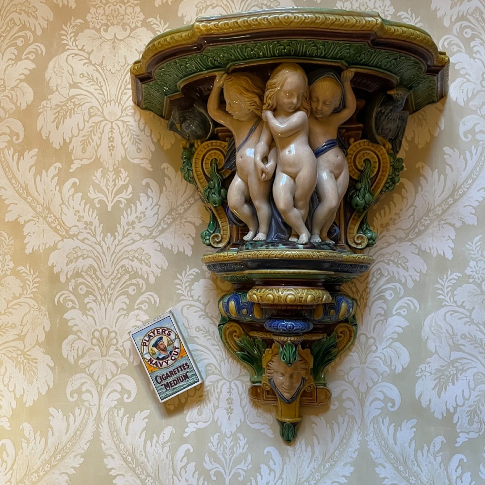 A Victorian Minton majolica wall bracket featuring three sculpted cherubs supporting a green glaze top shelf above a mascaron base.
