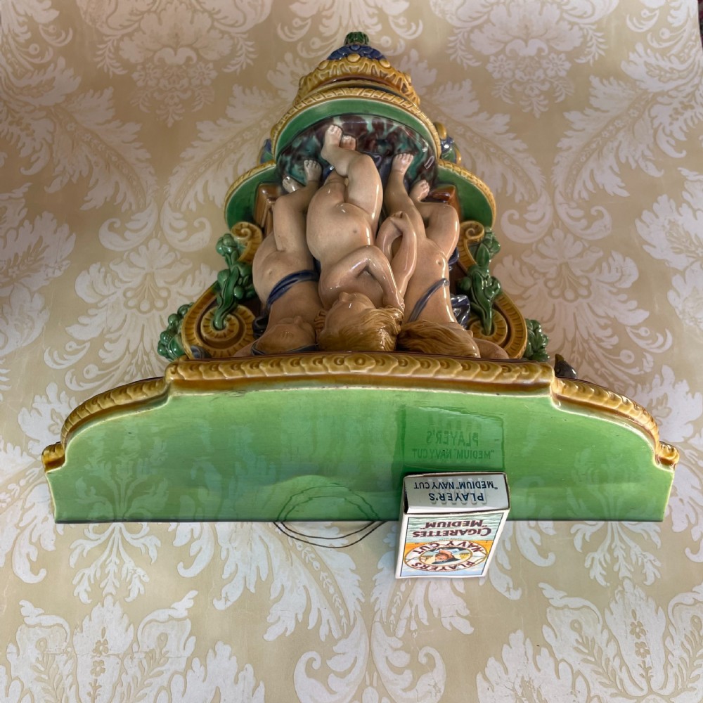 A Victorian Minton majolica wall bracket featuring three sculpted cherubs supporting a green glaze top shelf above a mascaron base. View II