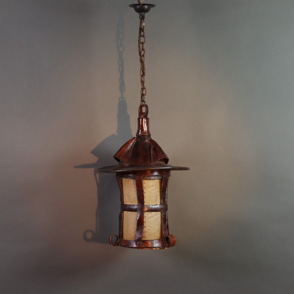 An Arts and Crafts ceiling lantern made of dark chestnut brown hand-stitched leather straps surrounding translucent vellum panels, suspended by a metal chain.