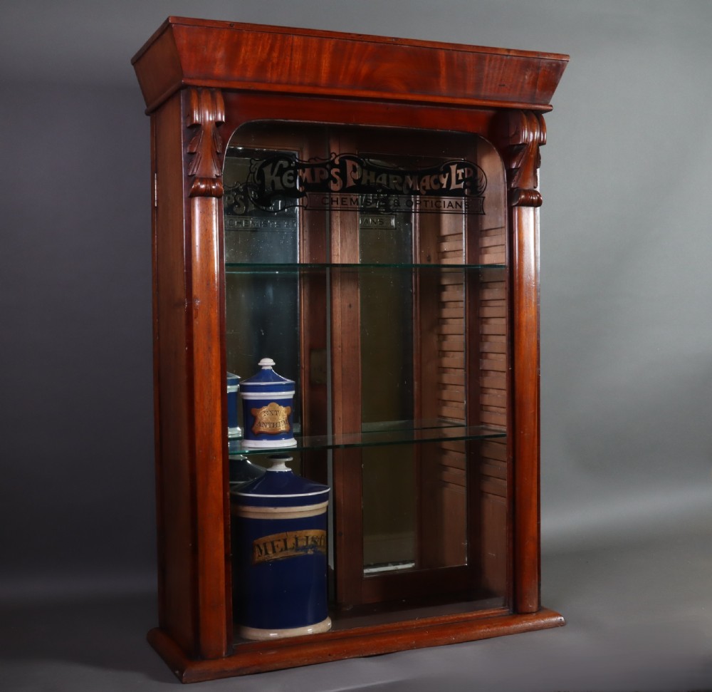 A Victorian mahogany pharmacy display cabinet featuring carved side columns, glass shelves, and gilded glass lettering reading Kemps Pharmacy Chemist and Opticians. View IV