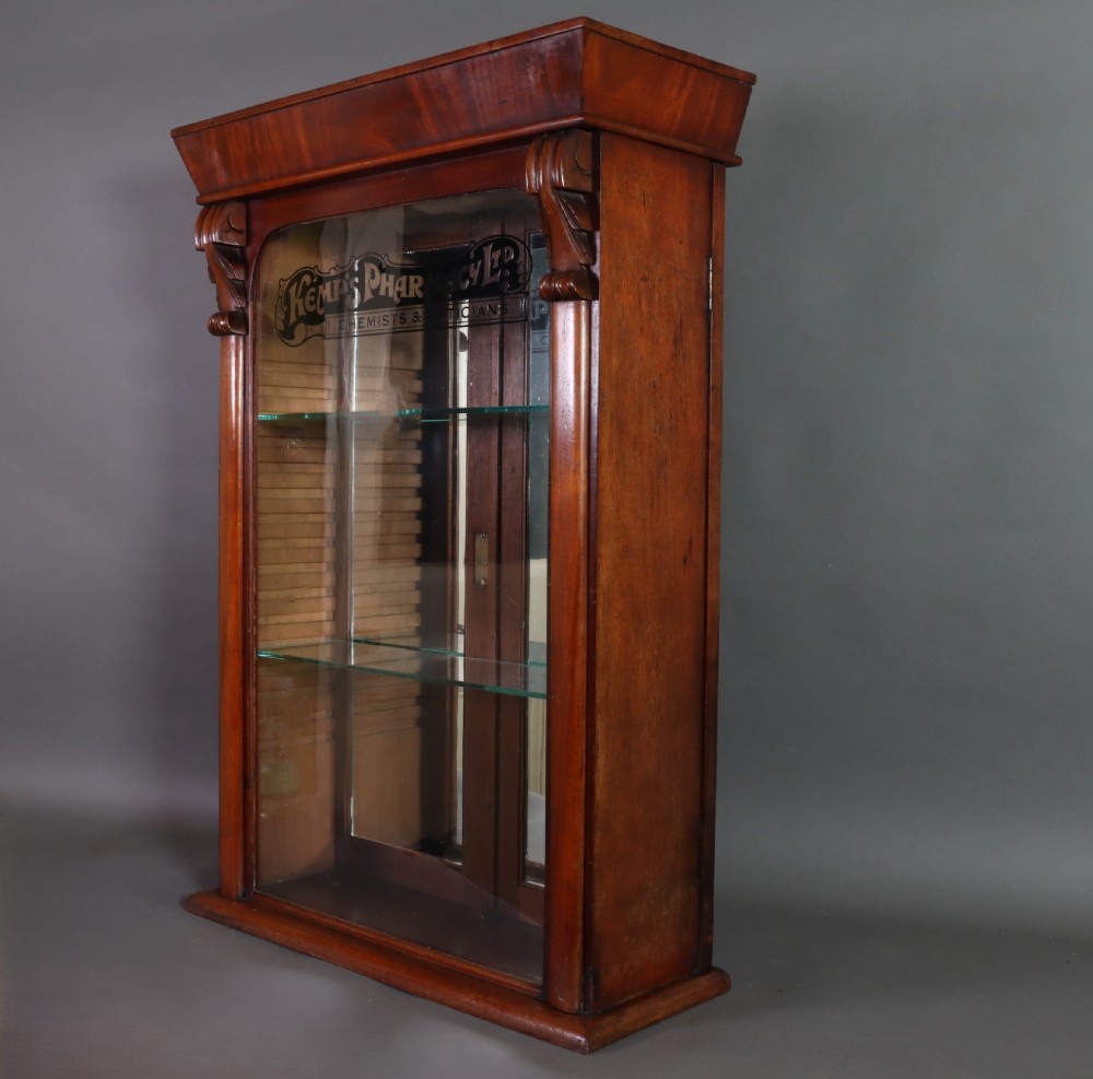 A Victorian mahogany pharmacy display cabinet featuring carved side columns, glass shelves, and gilded glass lettering reading Kemps Pharmacy Chemist and Opticians. View II