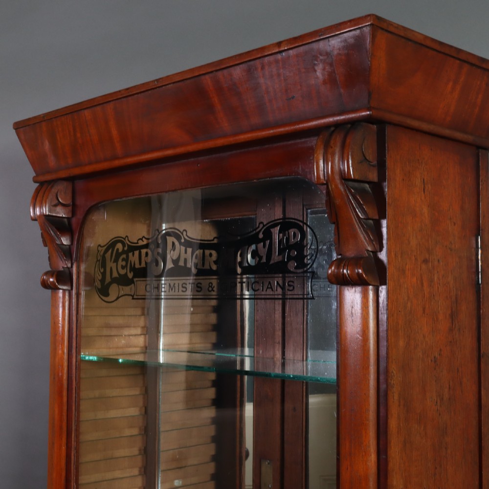 A Victorian mahogany pharmacy display cabinet featuring carved side columns, glass shelves, and gilded glass lettering reading Kemps Pharmacy Chemist and Opticians. View III