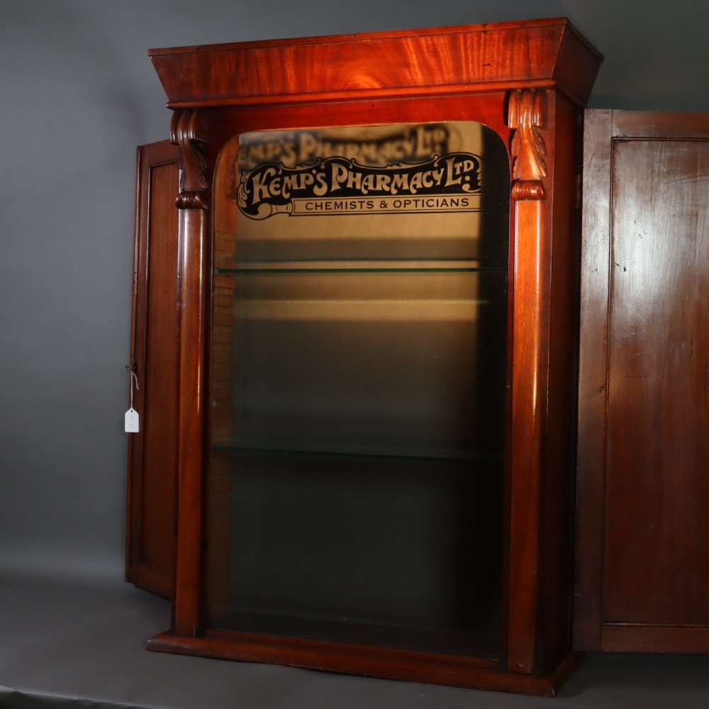 A Victorian mahogany pharmacy display cabinet featuring carved side columns, glass shelves, and gilded glass lettering reading Kemps Pharmacy Chemist and Opticians. View VI