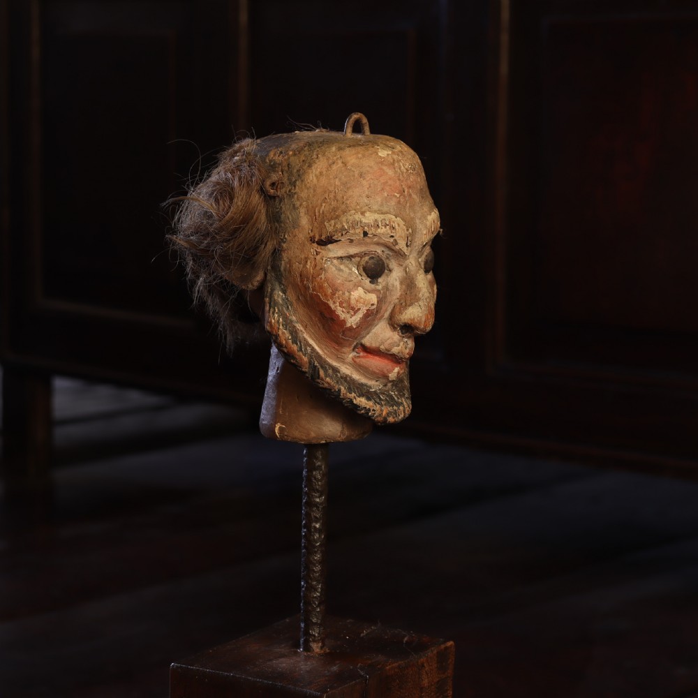 A nineteenth-century carved and polychrome painted wooden puppet head featuring natural hair and a bearded face, displayed on a metal stand.