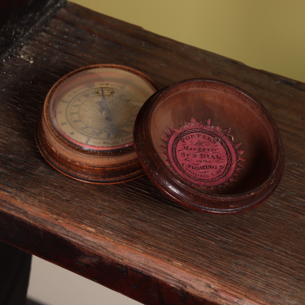 An open circular turned wood case displaying a Georgian magnetic pocket sundial with a pink printed makers label inside the lid.