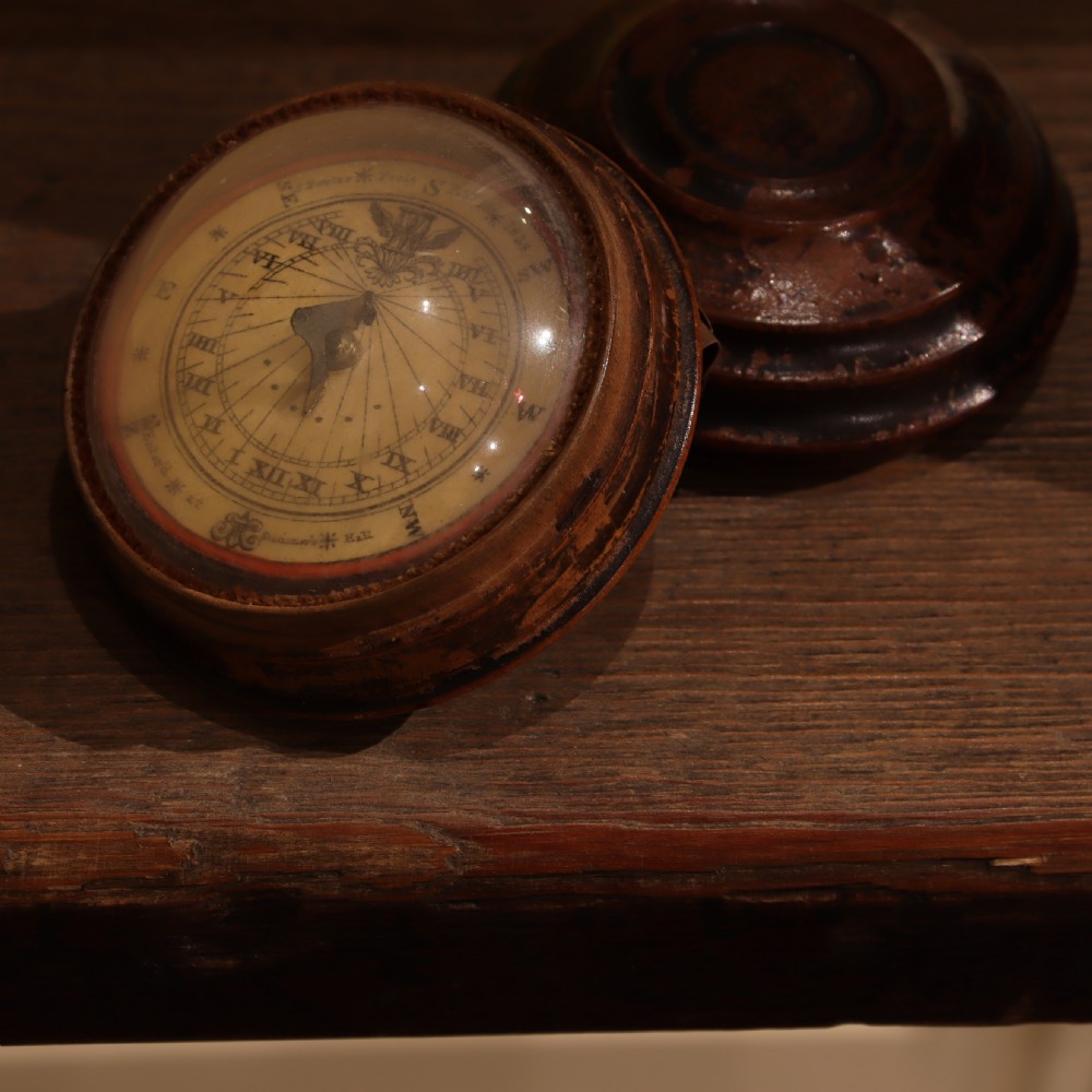 An open circular turned wood case displaying a Georgian magnetic pocket sundial with a pink printed makers label inside the lid. View II