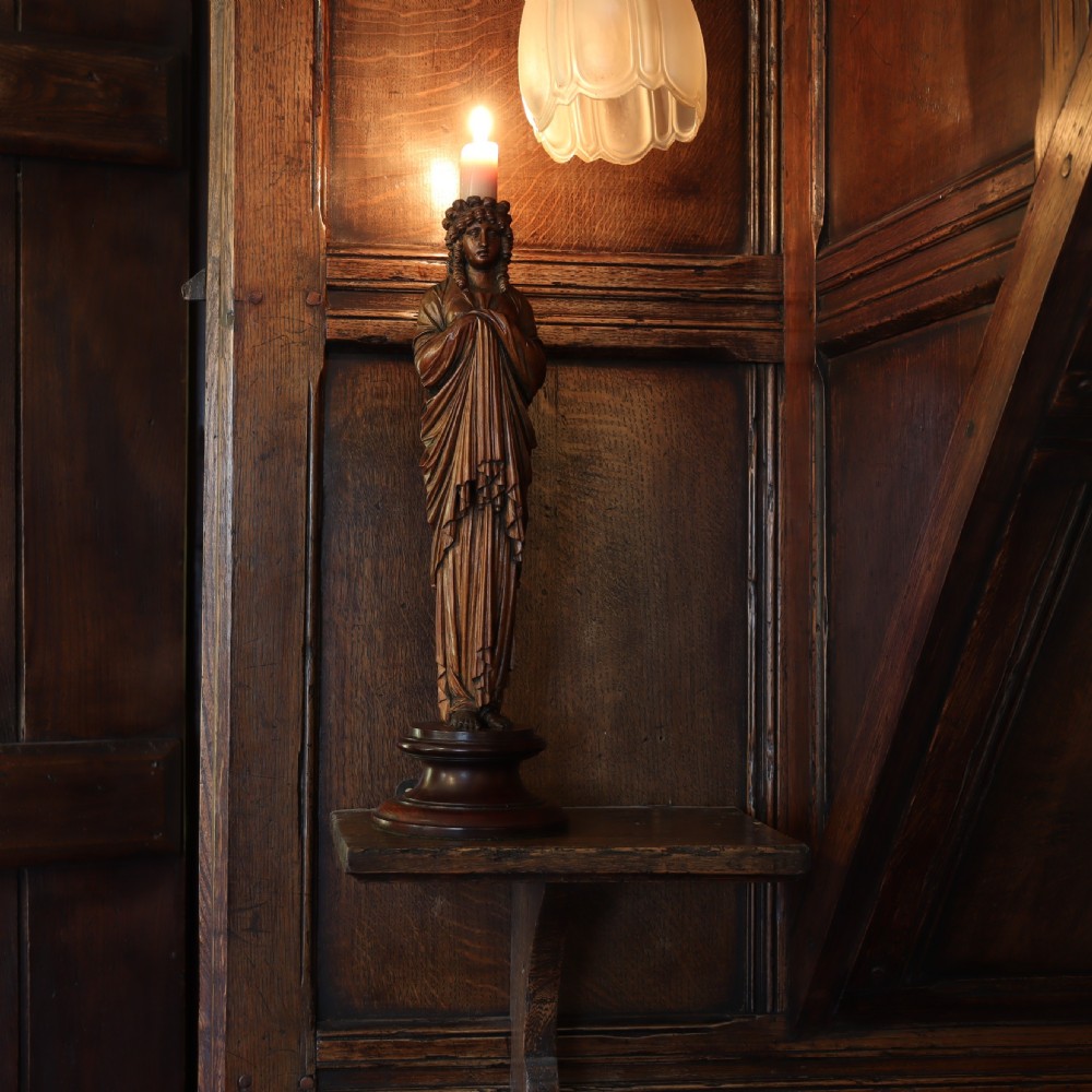 A 19th-century carved wooden candlestick shaped as a draped female figure standing on a round turned socle base. View II