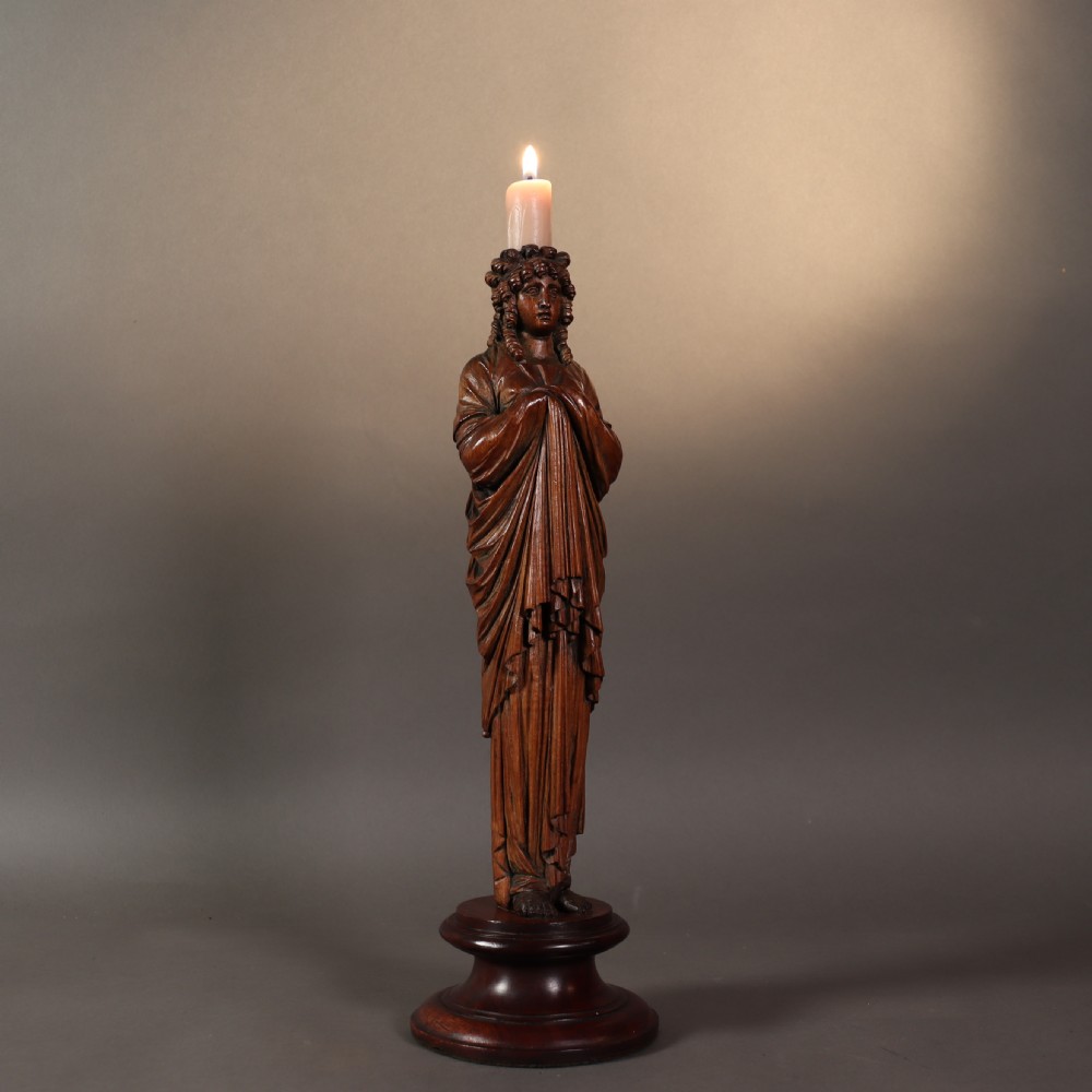 A 19th-century carved wooden candlestick shaped as a draped female figure standing on a round turned socle base.