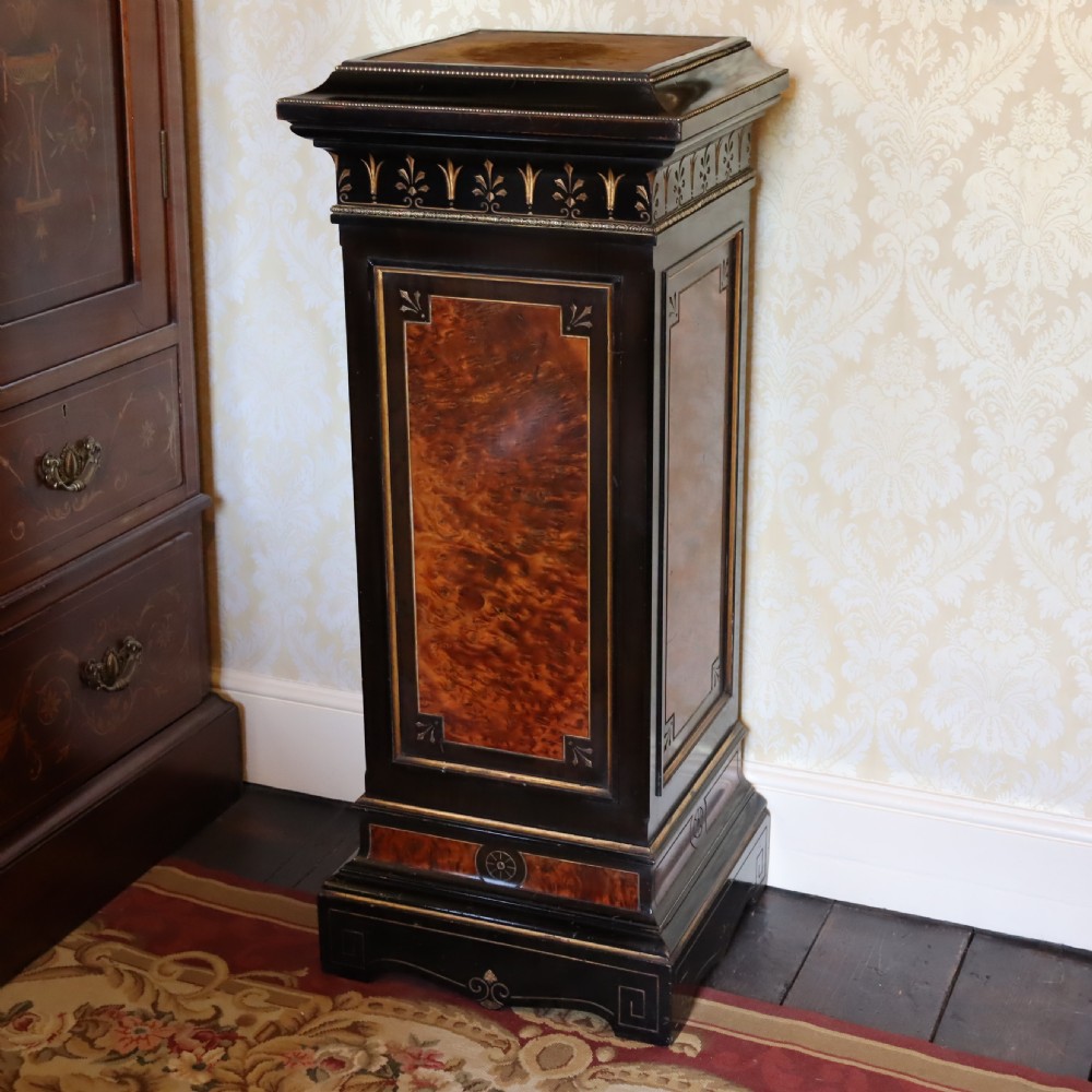 An ebonised Victorian Aesthetic Movement display pedestal featuring inset burr walnut panels, gilt incised frieze detailing, and a moulded square top.