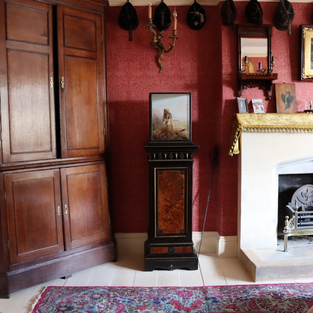 An ebonised Victorian Aesthetic Movement display pedestal featuring inset burr walnut panels, gilt incised frieze detailing, and a moulded square top. View X