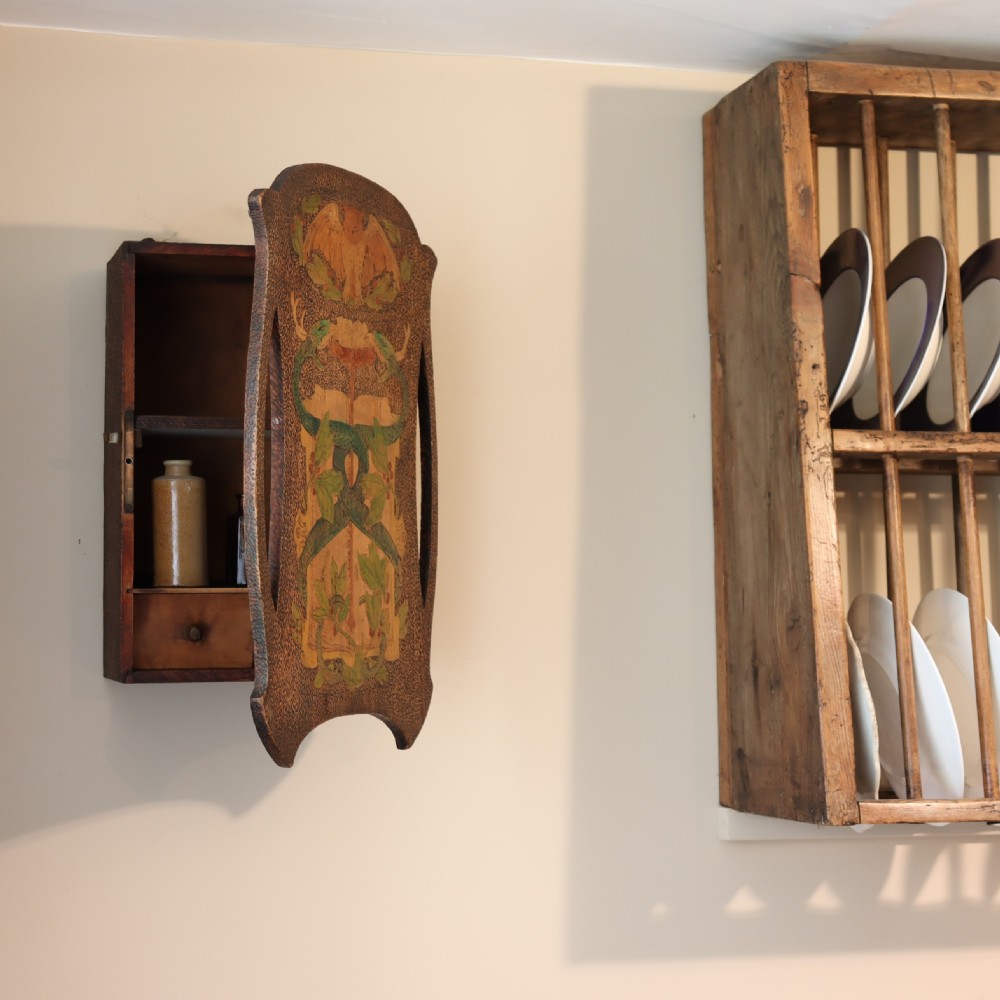 An Arts and Crafts wooden wall cupboard decorated with pyrography and painted motifs depicting an owl, green serpents, and foliage against a dark textured surface. View II