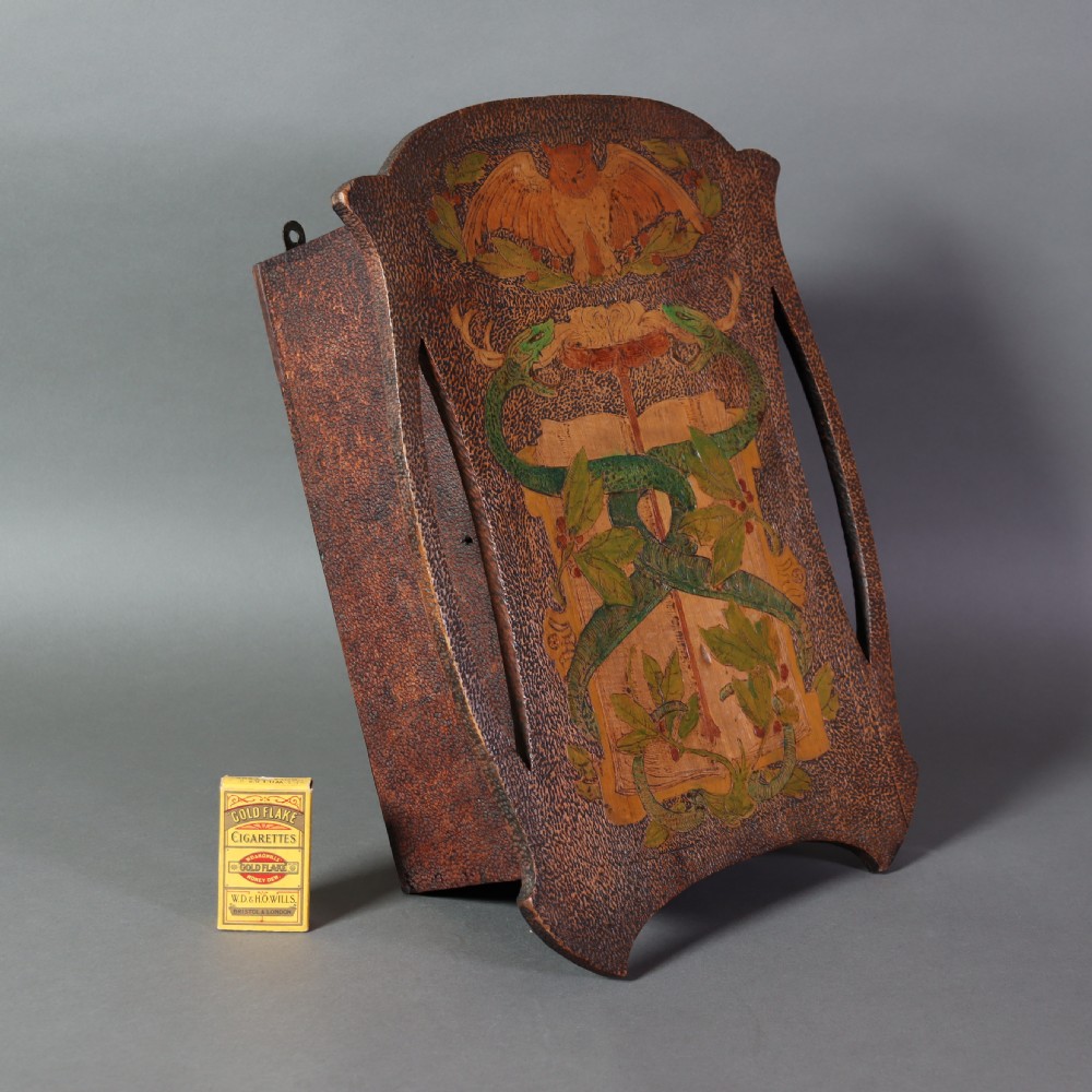An Arts and Crafts wooden wall cupboard decorated with pyrography and painted motifs depicting an owl, green serpents, and foliage against a dark textured surface. View V