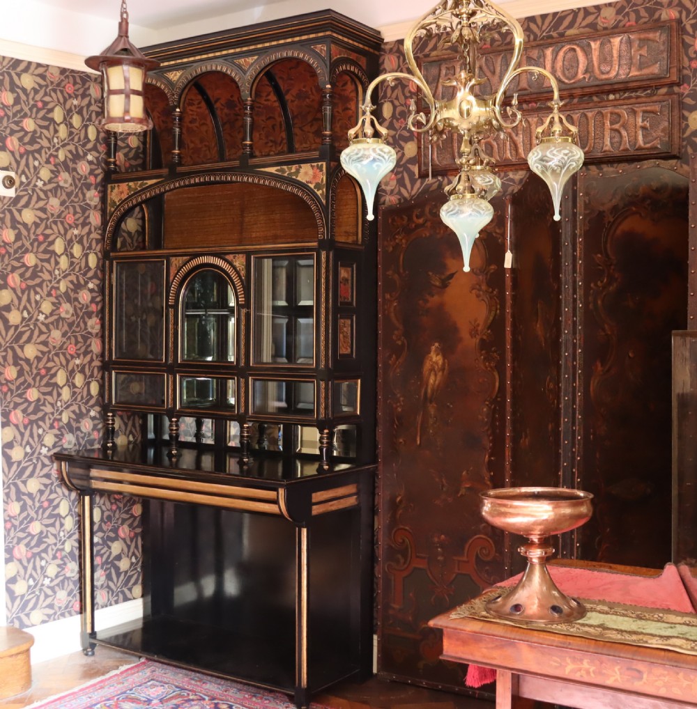 An Aesthetic Movement ebonised mahogany display cabinet featuring gilt highlights, turned spindle arcades, painted floral panels, and mirrored doors.