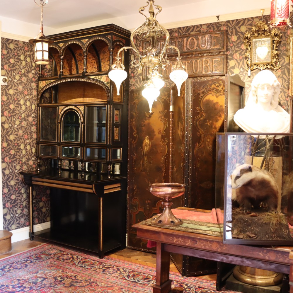 An Aesthetic Movement ebonised mahogany display cabinet featuring gilt highlights, turned spindle arcades, painted floral panels, and mirrored doors. View II
