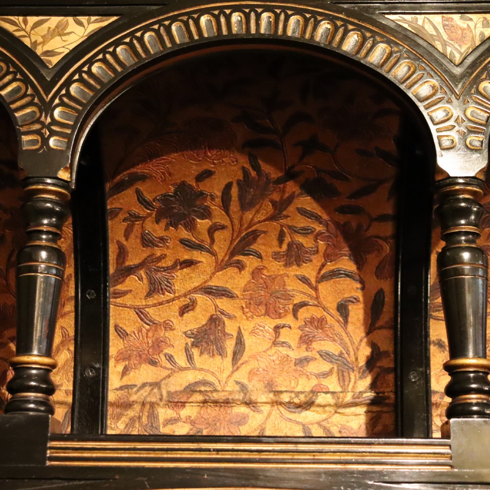 An Aesthetic Movement ebonised mahogany display cabinet featuring gilt highlights, turned spindle arcades, painted floral panels, and mirrored doors. View IV