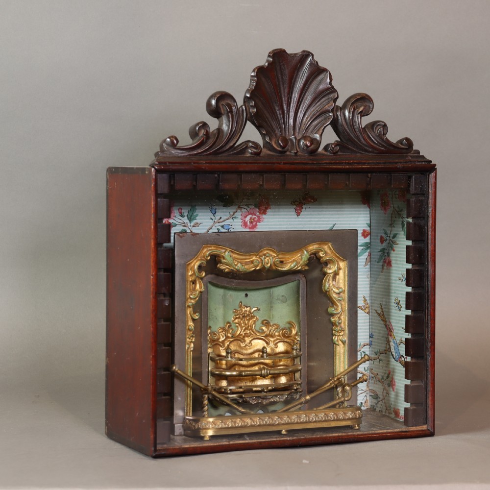 A Victorian miniature fireplace model with brass and iron hearth details, enclosed in a dark wood cabinet with a shell carved pediment.