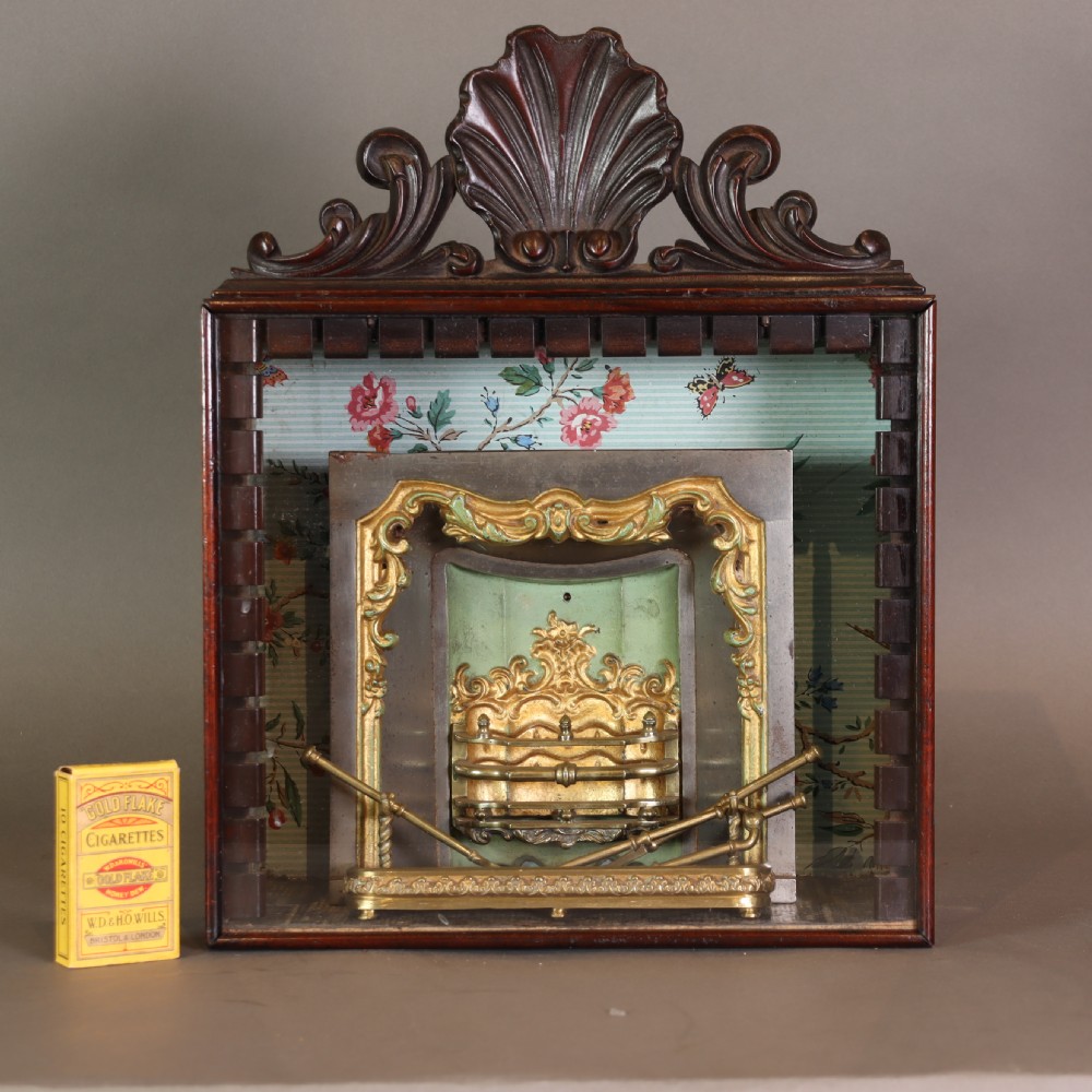 A Victorian miniature fireplace model with brass and iron hearth details, enclosed in a dark wood cabinet with a shell carved pediment. View IV
