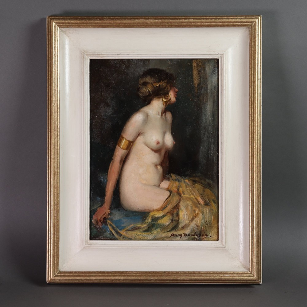 An oil painting on board depicting a seated female nude in profile wearing gold jewellery, presented in a painted cream and gold wood frame.