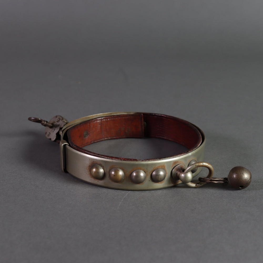 An Edwardian white metal dog collar featuring a brown leather lining, raised metal studs, an attached brass bell, and a functional padlock with key.