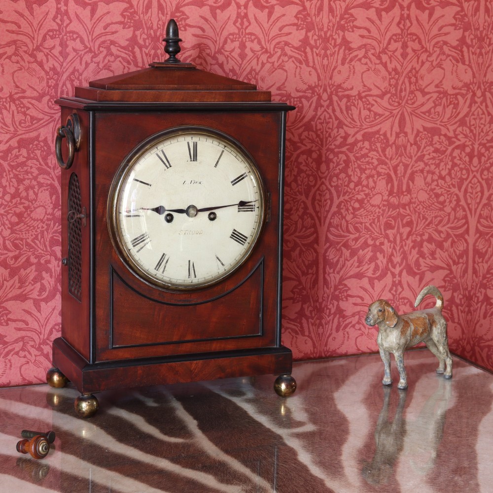 A George IV mahogany bracket clock featuring ebony inlaid lines, brass ball feet, side carrying handles, and a circular white dial signed by L. Vick of Stroud.