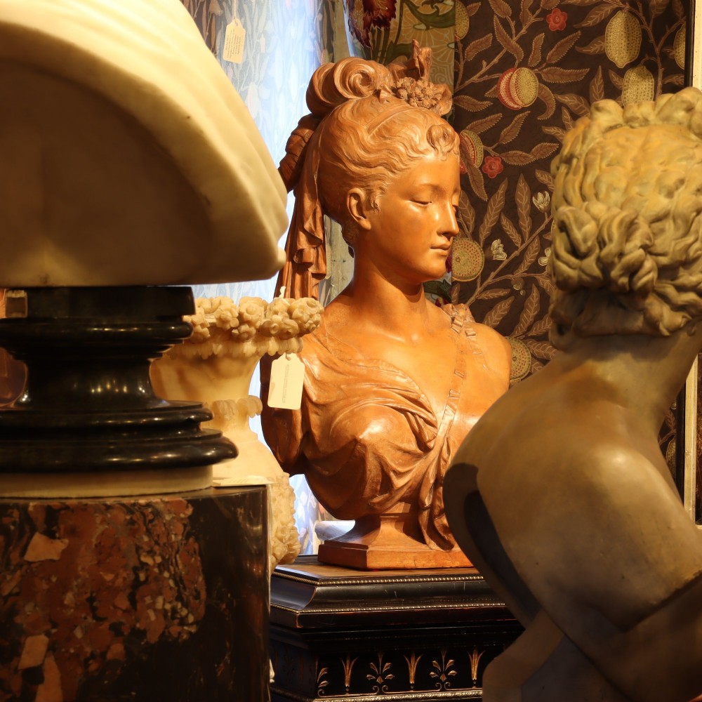 A Neoclassical terracotta bust depicting a young woman with a floral hair arrangement and draped semi-nude torso set on an integrated base. View IV