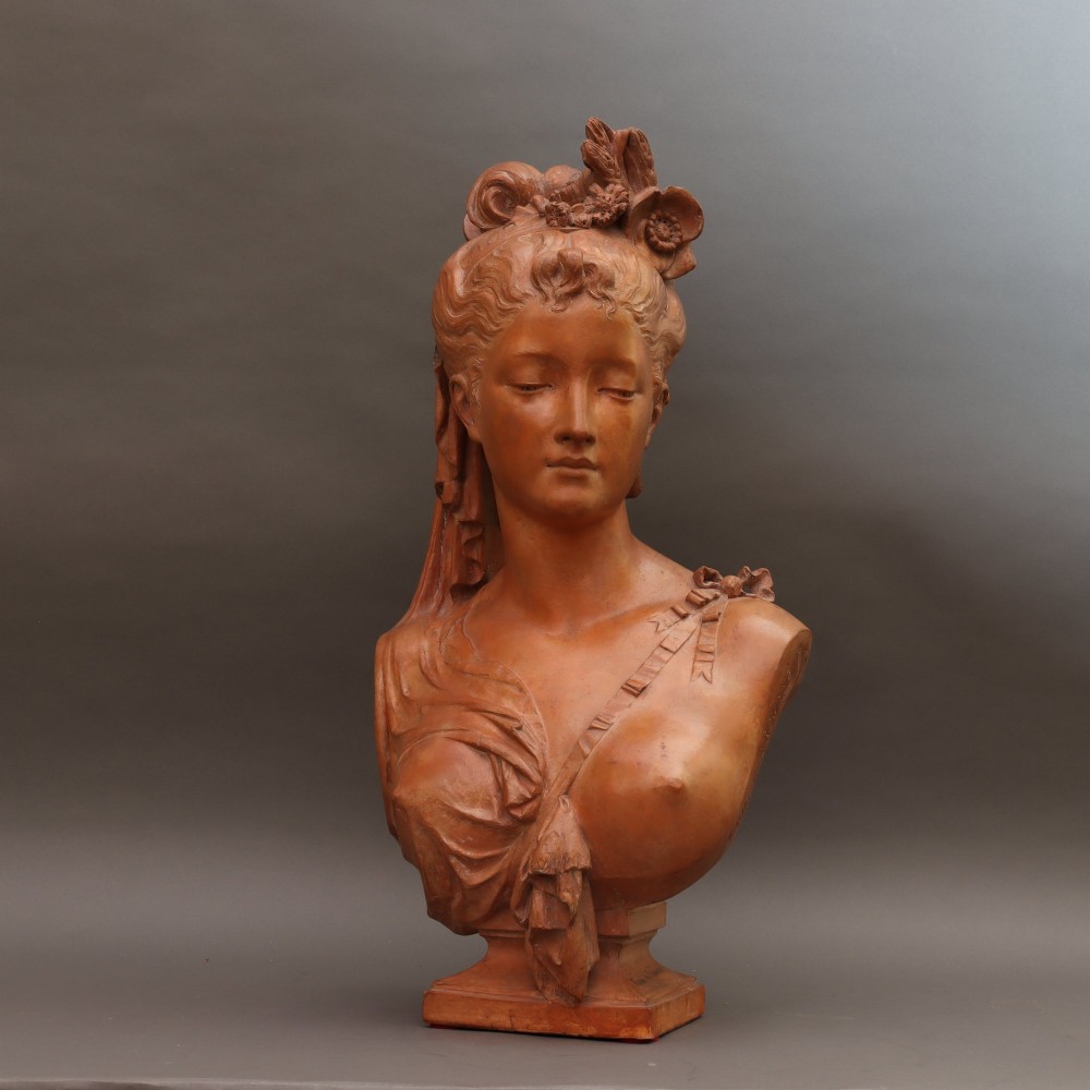 A Neoclassical terracotta bust depicting a young woman with a floral hair arrangement and draped semi-nude torso set on an integrated base. View V