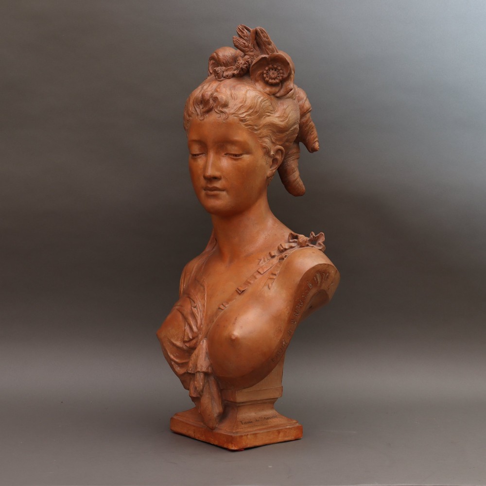 A Neoclassical terracotta bust depicting a young woman with a floral hair arrangement and draped semi-nude torso set on an integrated base.