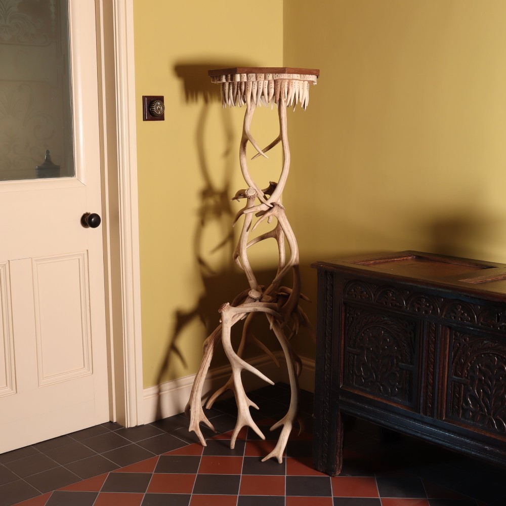 A tall, rustic torchiere stand constructed from intertwined natural deer antlers supporting an octagonal wooden top trimmed with downward-pointing antler tines.