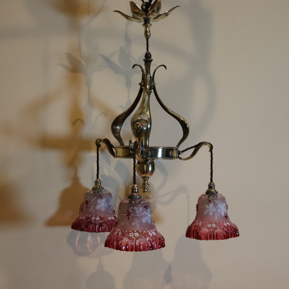 An Arts and Crafts brass ceiling light featuring three curved arms holding ruffled cranberry glass shades with painted floral motifs.