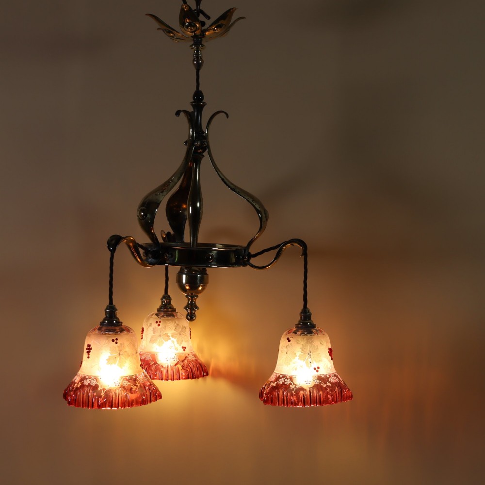 An Arts and Crafts brass ceiling light featuring three curved arms holding ruffled cranberry glass shades with painted floral motifs. View III