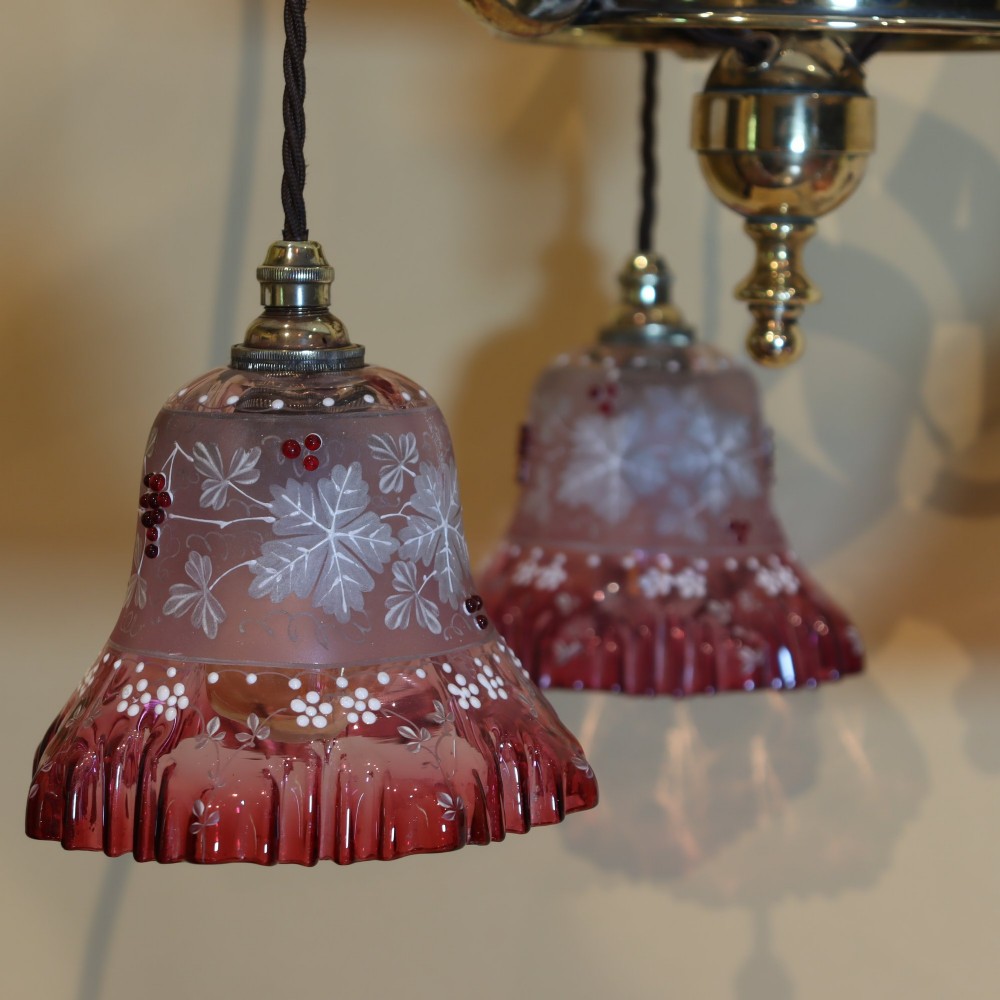An Arts and Crafts brass ceiling light featuring three curved arms holding ruffled cranberry glass shades with painted floral motifs. View V