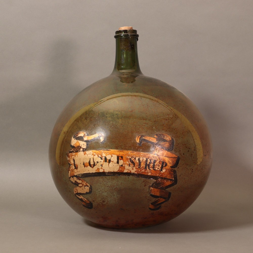 A large spherical olive green glass apothecary carboy featuring a painted banner with gilded scroll lettering. View II