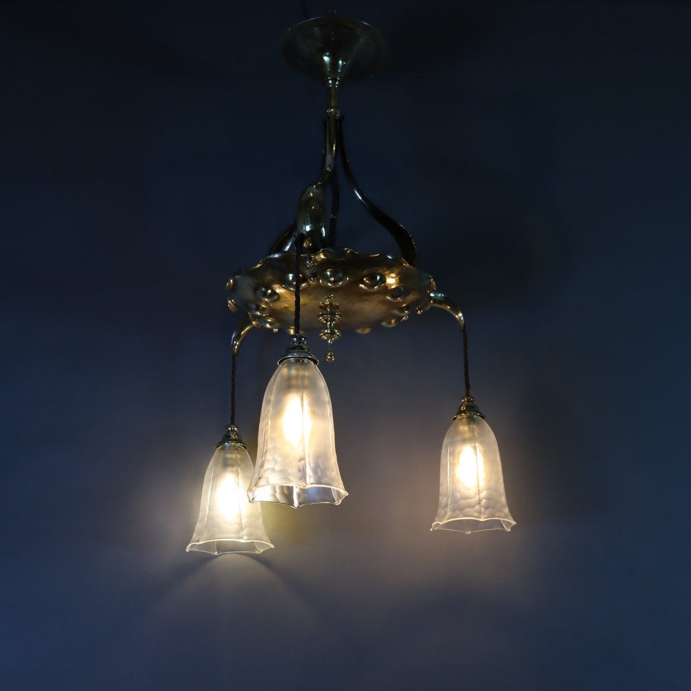 An antique Arts and Crafts brass ceiling fixture featuring a central hand-hammered canopy and three suspended drop pendant lights with frosted bell glass shades. View VIII