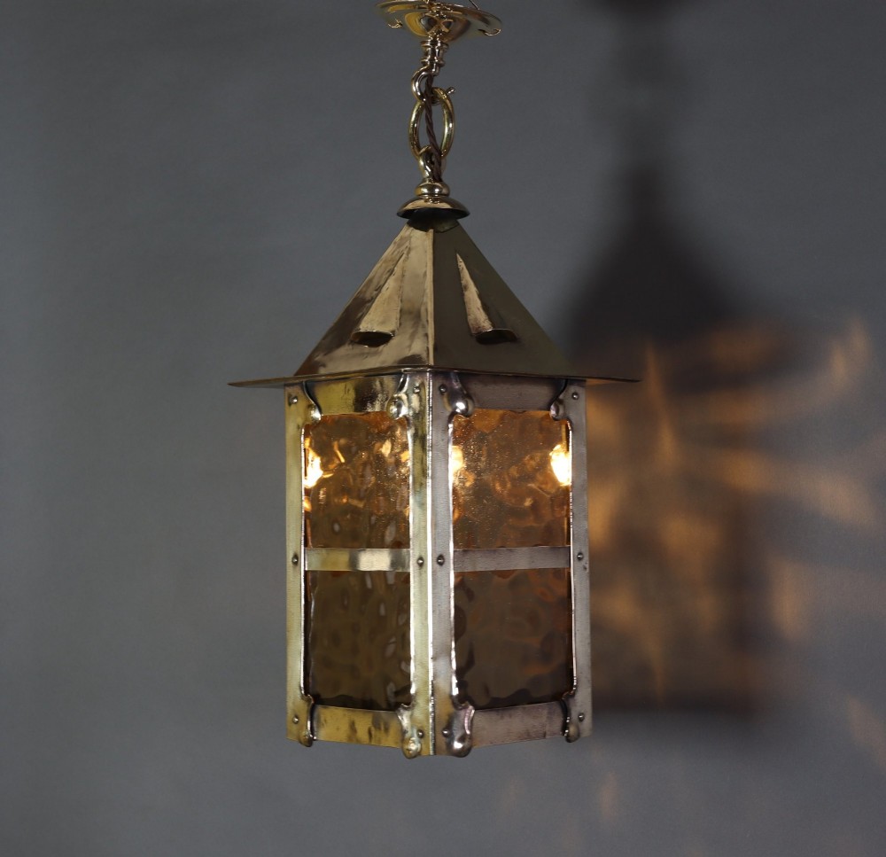 An Arts and Crafts style brass hanging lantern featuring amber textured glass panes, a riveted square body, a pyramidal top hood, and a ceiling chain suspension.