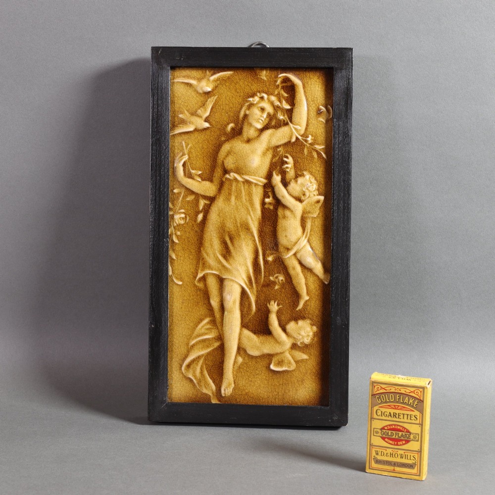 A rectangular moulded relief ceramic tile depicting an allegorical maiden with cherubs and birds, finished in an amber translucent glaze and set in a simple dark wooden frame. View IV