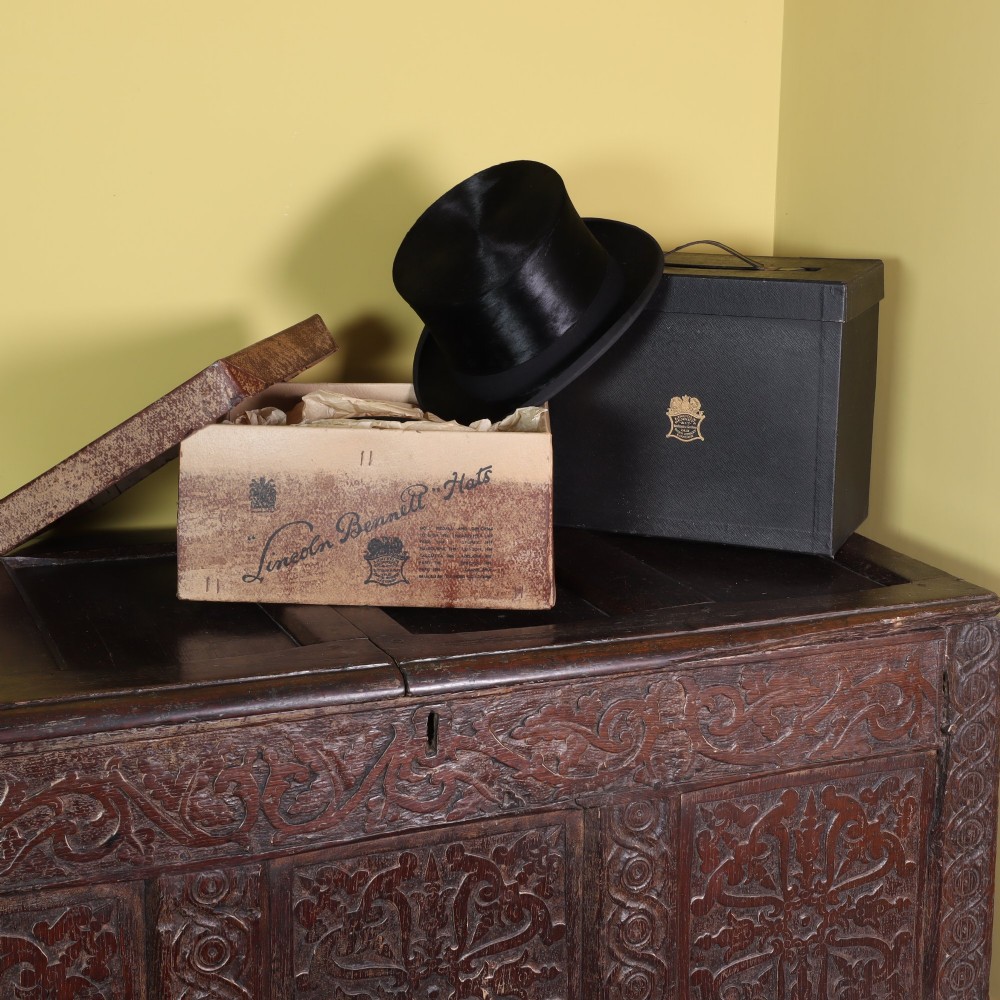 A black silk top hat resting on stacked fitted hat boxes beside a black bowler hat by Lincoln Bennett of London. View II