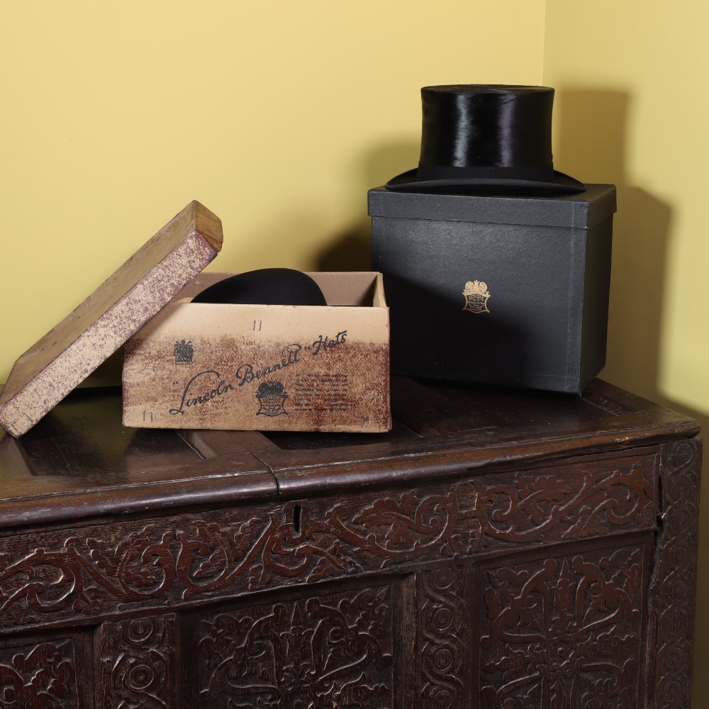 A black silk top hat resting on stacked fitted hat boxes beside a black bowler hat by Lincoln Bennett of London. View IV