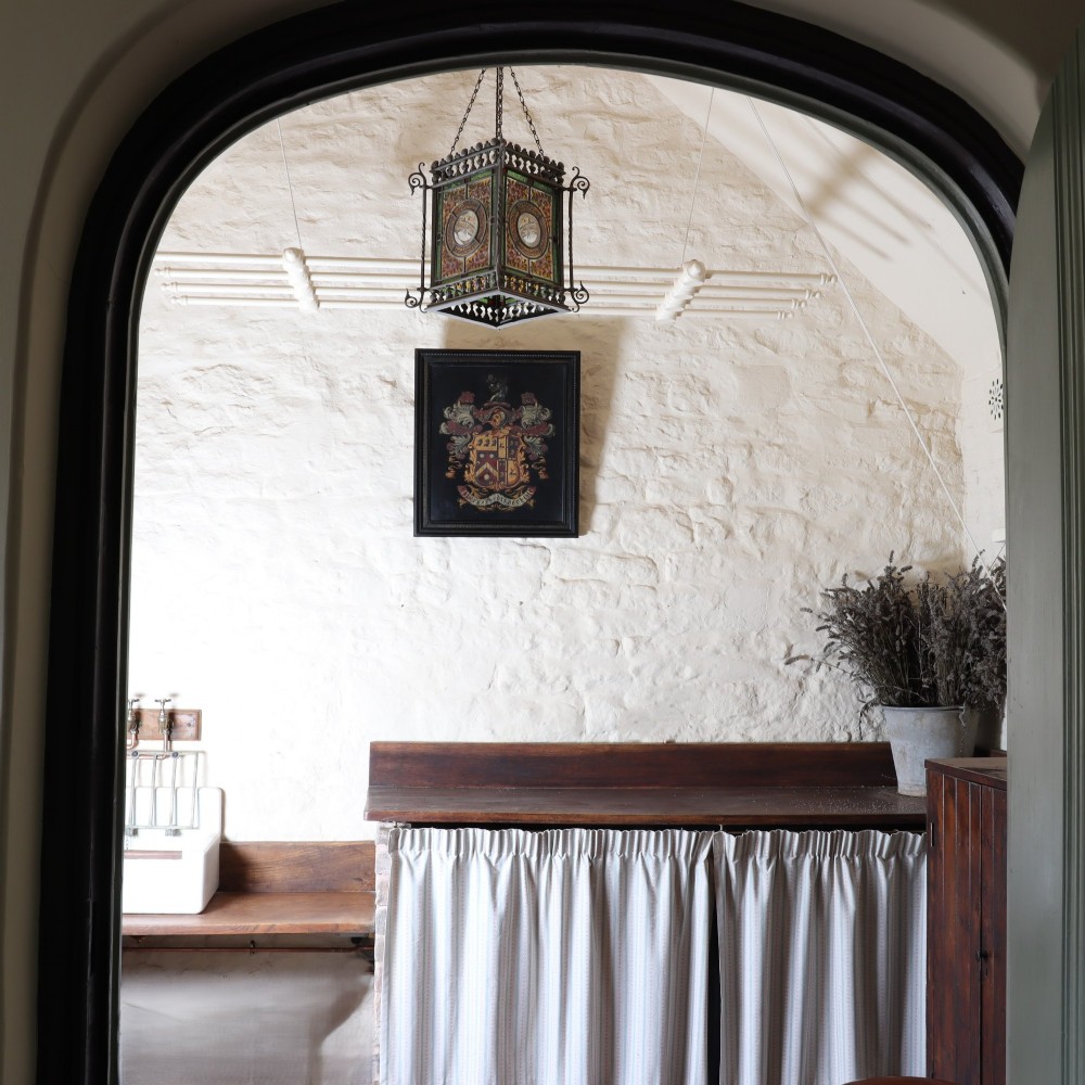 A large black-framed 19th-century panel featuring a painted coat of arms with red and cream mantling above a heraldic motto scroll. View V