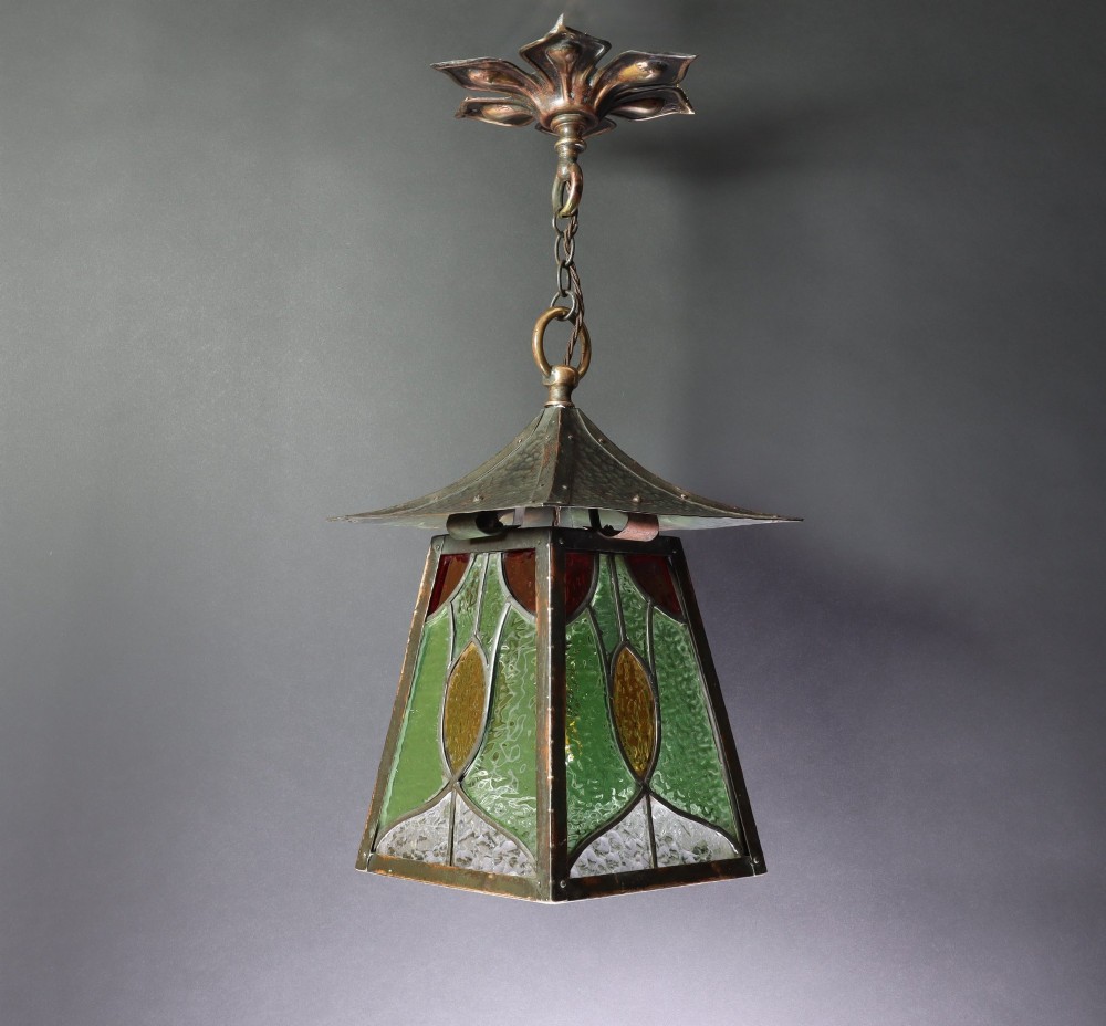An Arts and Crafts planished copper hanging lantern featuring leaded green, yellow, and clear glass panels suspended from a chain and floral ceiling rose.