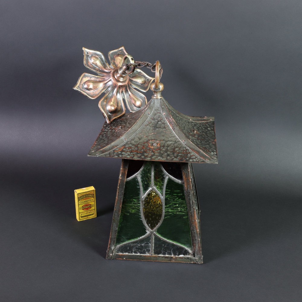 An Arts and Crafts planished copper hanging lantern featuring leaded green, yellow, and clear glass panels suspended from a chain and floral ceiling rose. View V