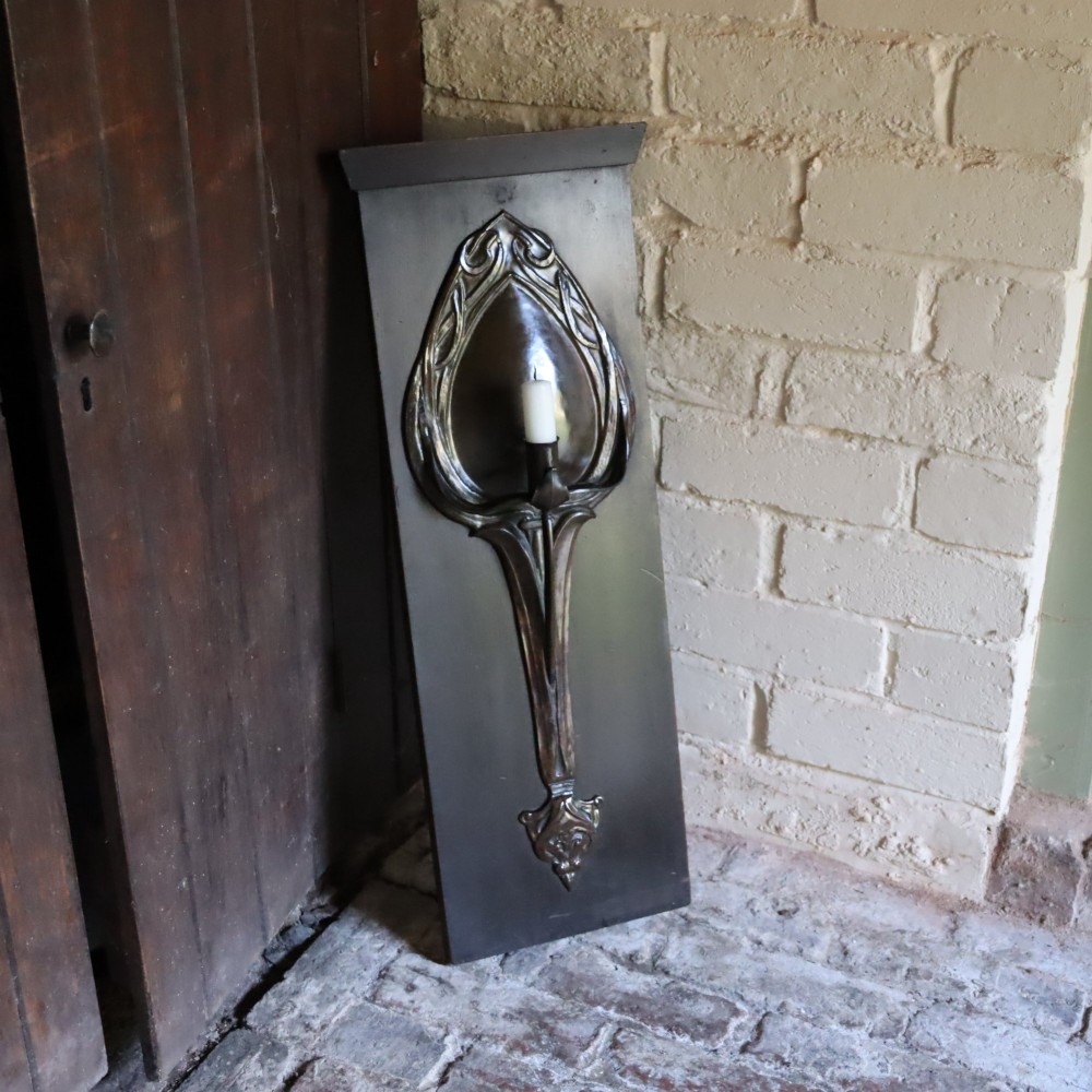 A patinated copper wall sconce with stylized Art Nouveau repousse details mounted on a dark rectangular wooden backboard. View III