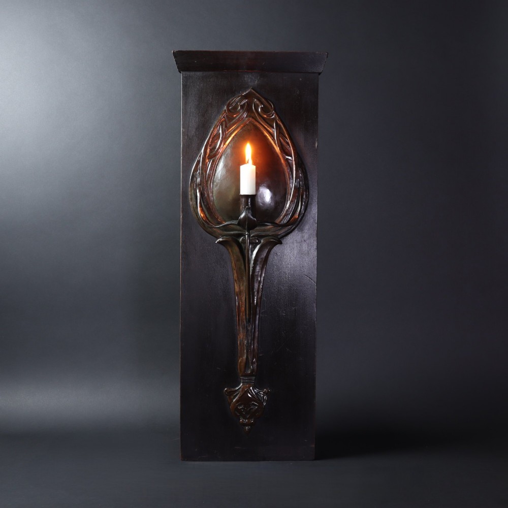 A patinated copper wall sconce with stylized Art Nouveau repousse details mounted on a dark rectangular wooden backboard. View IV