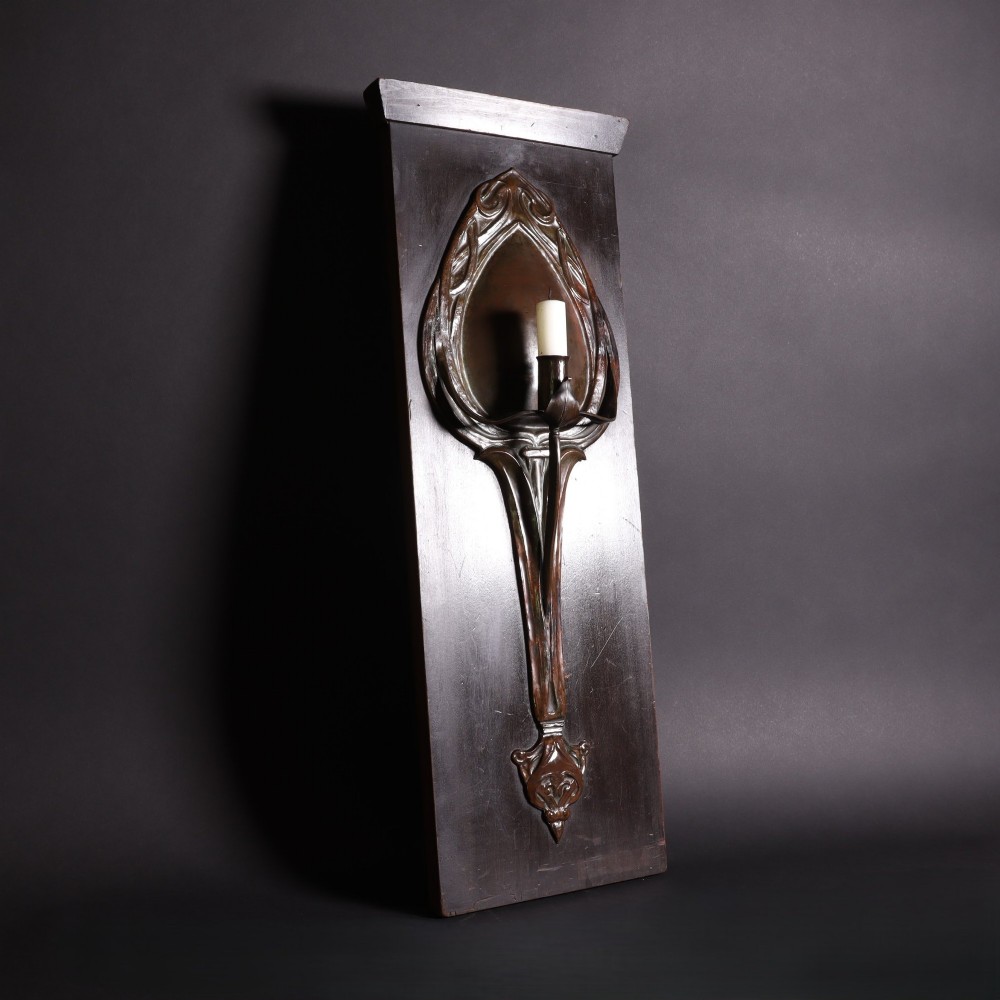 A patinated copper wall sconce with stylized Art Nouveau repousse details mounted on a dark rectangular wooden backboard. View V