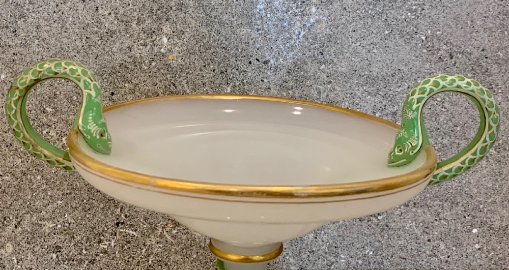 An antique white opaline glass centrepiece bowl featuring hand-painted green and gold serpent handles and a coiled snake motif around the pedestal base, resting on a transparent stand. View VI