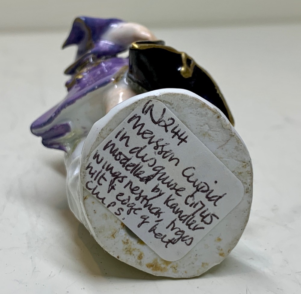 A small Meissen porcelain figurine of winged Cupid disguised in an 18th-century purple coat and powdered wig, resting on a sculpted rocky base. View IX