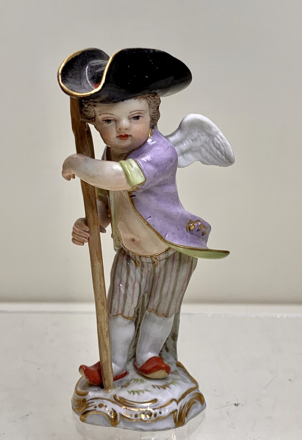 An 18th-century Meissen porcelain figurine depicting a winged Cupid disguised in a black tricorn hat, purple coat, and striped breeches resting on a staff.