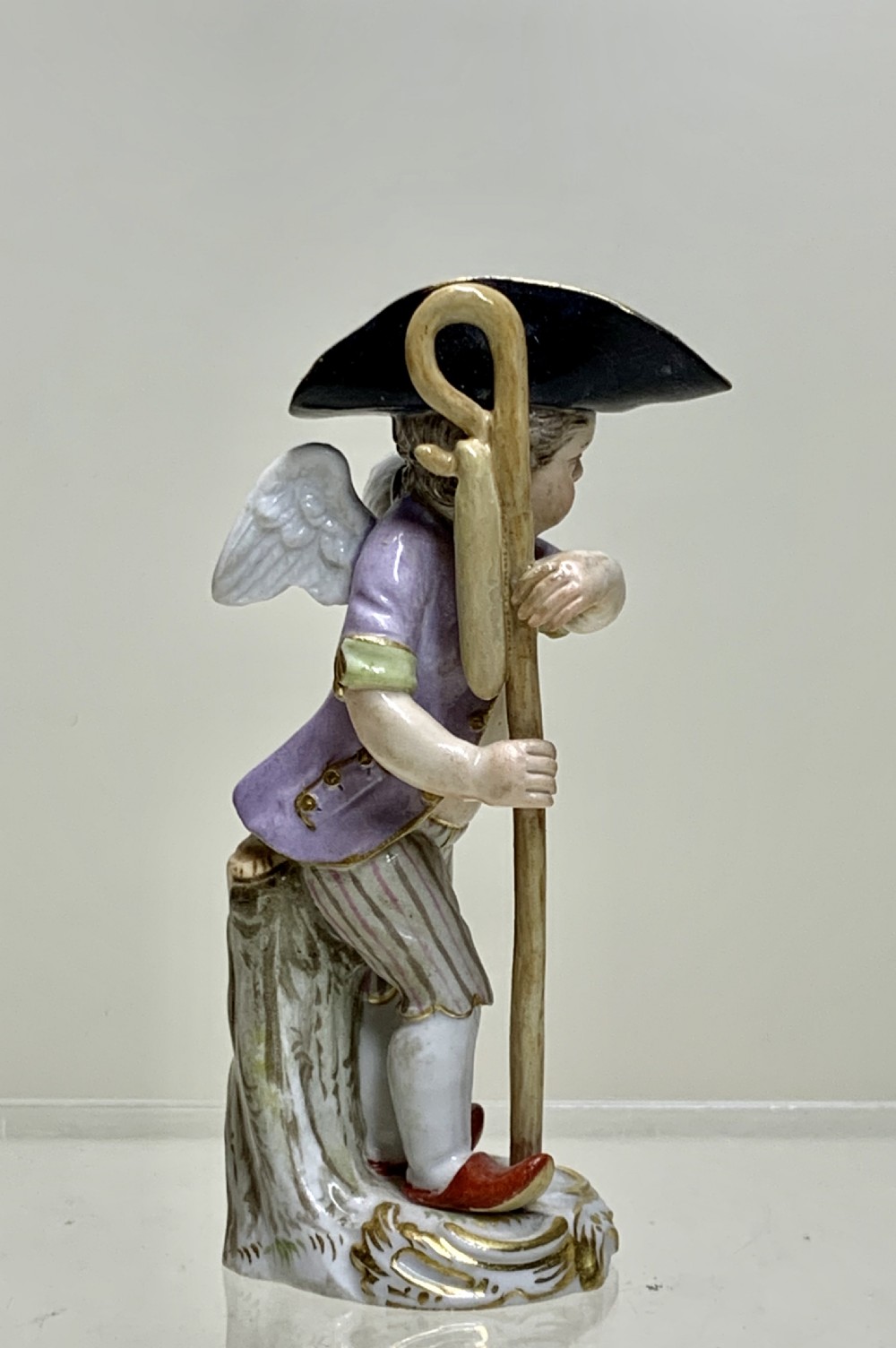 An 18th-century Meissen porcelain figurine depicting a winged Cupid disguised in a black tricorn hat, purple coat, and striped breeches resting on a staff. View III